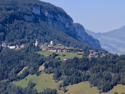 A distant landscape view of a small village nestled on a green hillside at the foot of a steep, forested limestone cliff face, featuring a prominent church tower and several large buildings.