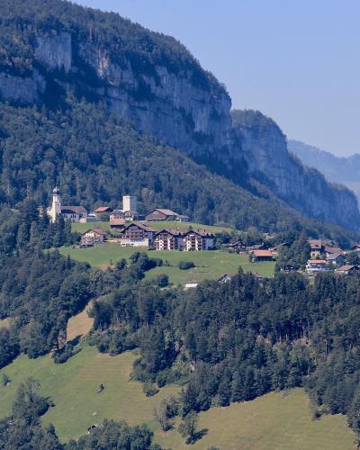 A distant landscape view of a small village nestled on a green hillside at the foot of a steep, forested limestone cliff face, featuring a prominent church tower and several large buildings.