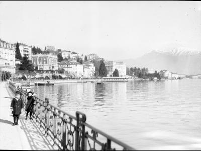 A black and white historical photograph showing two young boys standing on a paved promenade next to an ornate metal railing, overlooking a large lake with a city shoreline and mountains in the background.