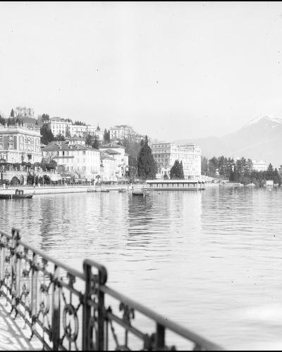 A black and white historical photograph showing two young boys standing on a paved promenade next to an ornate metal railing, overlooking a large lake with a city shoreline and mountains in the background.