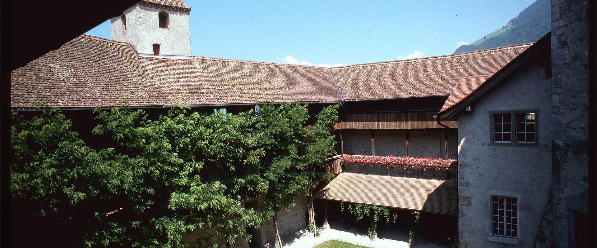 A view of a stone castle courtyard with a tiled roof, a small tower, green trees, and a grassy lawn under a blue sky.