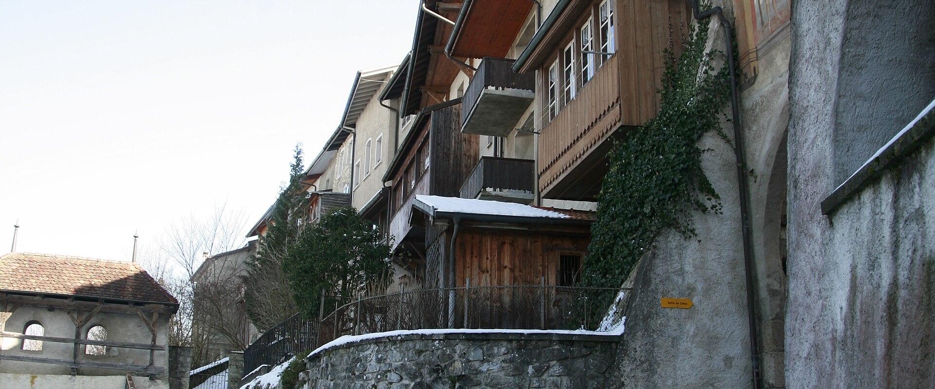 A snowy, narrow street in a historic European town, featuring traditional buildings with wooden balconies and a stone retaining wall, with a covered bridge visible in the background.