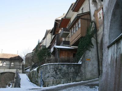 A snowy, narrow street in a historic European town, featuring traditional buildings with wooden balconies and a stone retaining wall, with a covered bridge visible in the background.