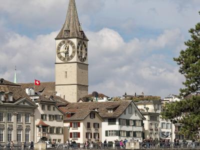 The image shows the St. Peter Church tower in Zurich, Switzerland, viewed from across the Limmat River with the Münsterbrücke bridge in the foreground; a Swiss flag is visible on a nearby building, and people are walking on the bridge.