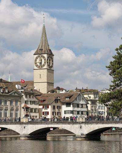 The image shows the St. Peter Church tower in Zurich, Switzerland, viewed from across the Limmat River with the Münsterbrücke bridge in the foreground; a Swiss flag is visible on a nearby building, and people are walking on the bridge.