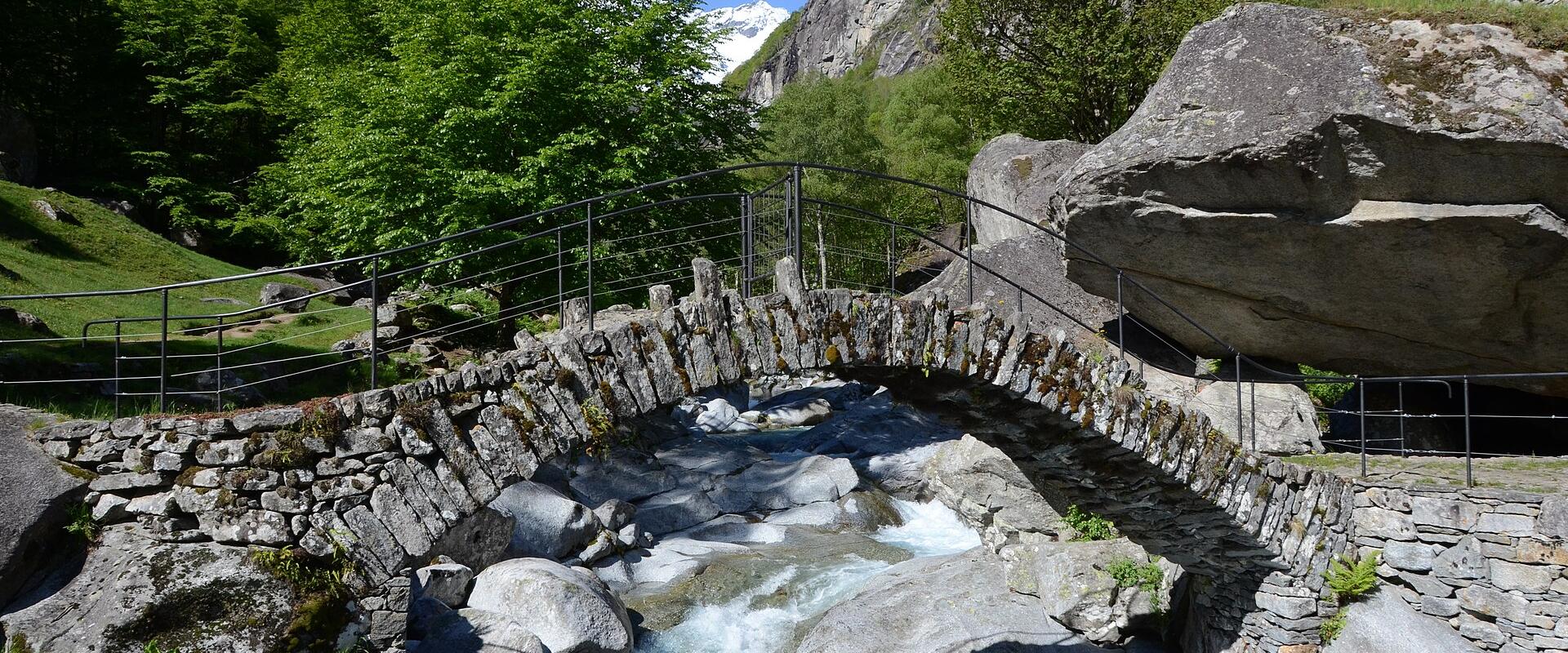 A traditional stone arch bridge with modern metal railings spanning a rocky stream in a green alpine valley with snow-capped mountains in the background.