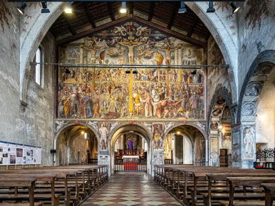 The interior of a church featuring a large, multi-scene fresco cycle on the back wall above the altar, wooden pews, and informational display boards along the left wall.