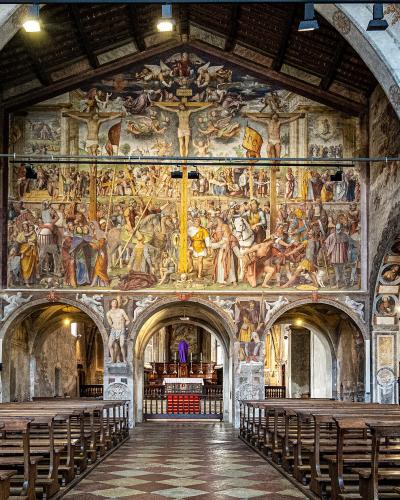 The interior of a church featuring a large, multi-scene fresco cycle on the back wall above the altar, wooden pews, and informational display boards along the left wall.