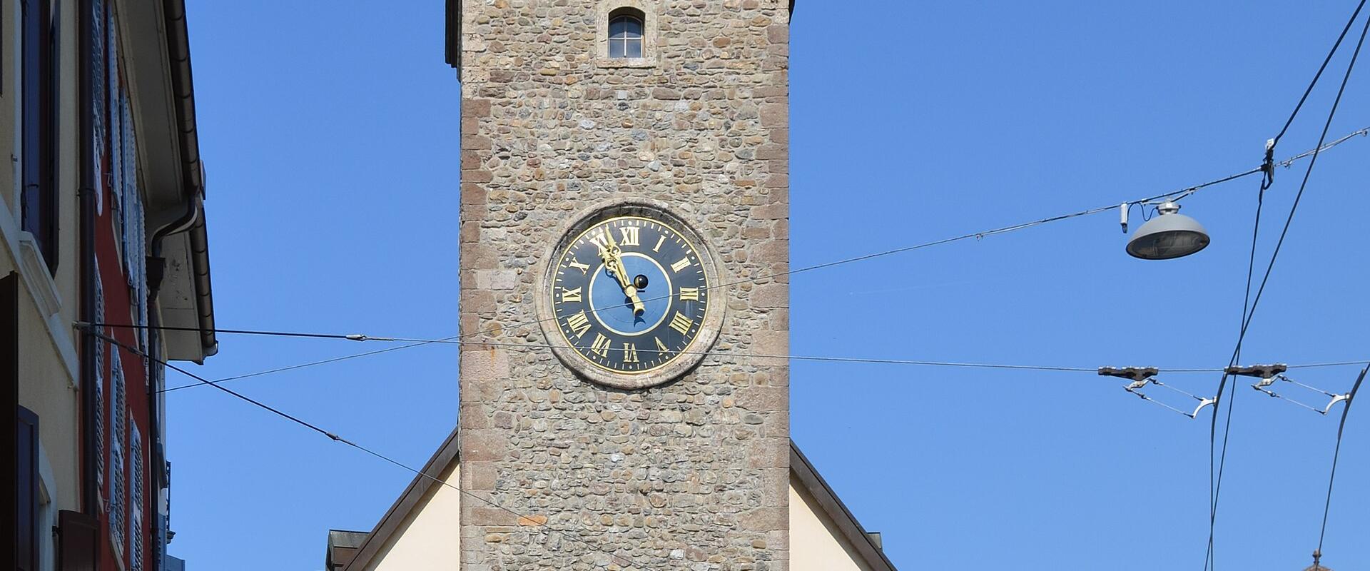 A tall, medieval stone clock tower with a green copper cupola and open belfry, situated in a European town square with surrounding buildings, a white van, and overhead tram lines.