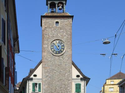 A tall, medieval stone clock tower with a green copper cupola and open belfry, situated in a European town square with surrounding buildings, a white van, and overhead tram lines.