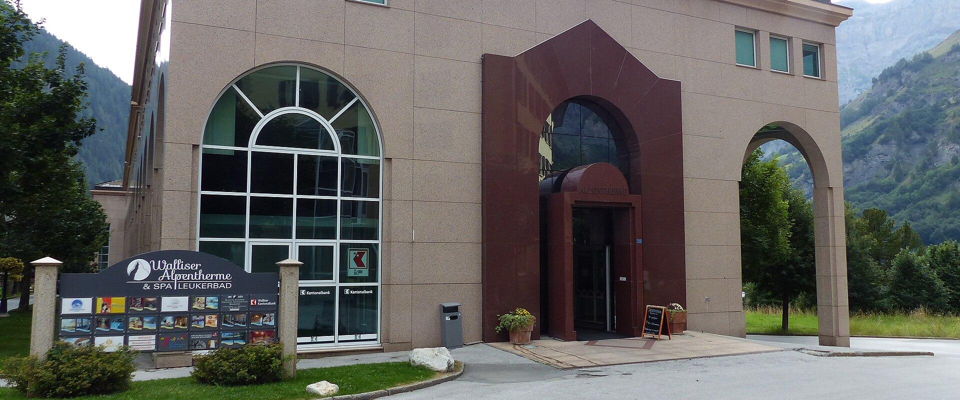 A modern beige building with large arched windows and a reddish-brown entrance archway, featuring a signboard in the foreground that reads "Walliser Alpentherme & SPA LEUKERBAD".