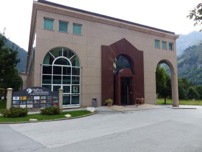 A modern beige building with large arched windows and a reddish-brown entrance archway, featuring a signboard in the foreground that reads "Walliser Alpentherme & SPA LEUKERBAD".