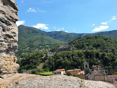A view from a stone wall looking out over a town with terracotta roofs, green hills, and a long fortified wall with towers running along the ridge.