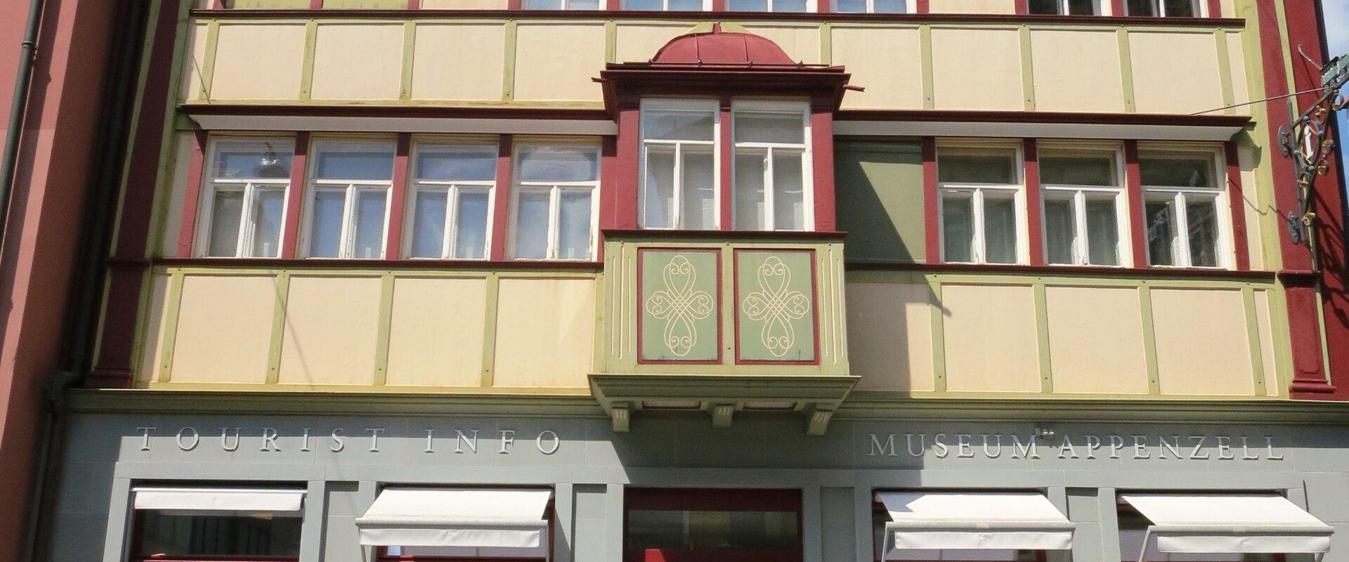 A multi-story building with traditional Appenzell architectural features, featuring a grey ground floor with signage reading "TOURIST INFO" and "MUSEUM APPENZELL", and a cream and red upper facade with a central bay window.