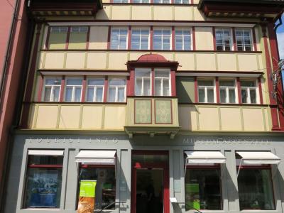 A multi-story building with traditional Appenzell architectural features, featuring a grey ground floor with signage reading "TOURIST INFO" and "MUSEUM APPENZELL", and a cream and red upper facade with a central bay window.