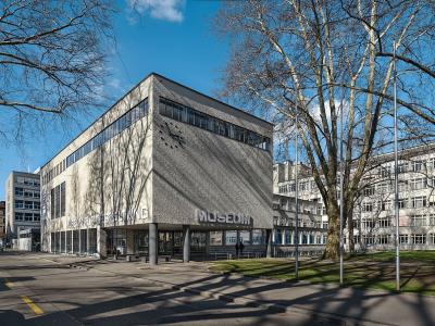 A modernist concrete building with large lettering reading "MUSEUM FÜR GESTALTUNG" and "MUSEUM" on its facade, set against a blue sky with bare trees and adjacent buildings.