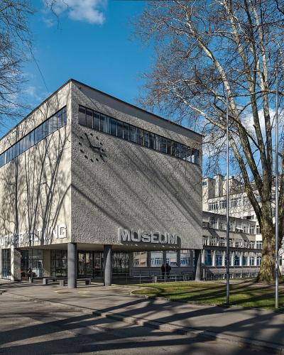 A modernist concrete building with large lettering reading "MUSEUM FÜR GESTALTUNG" and "MUSEUM" on its facade, set against a blue sky with bare trees and adjacent buildings.