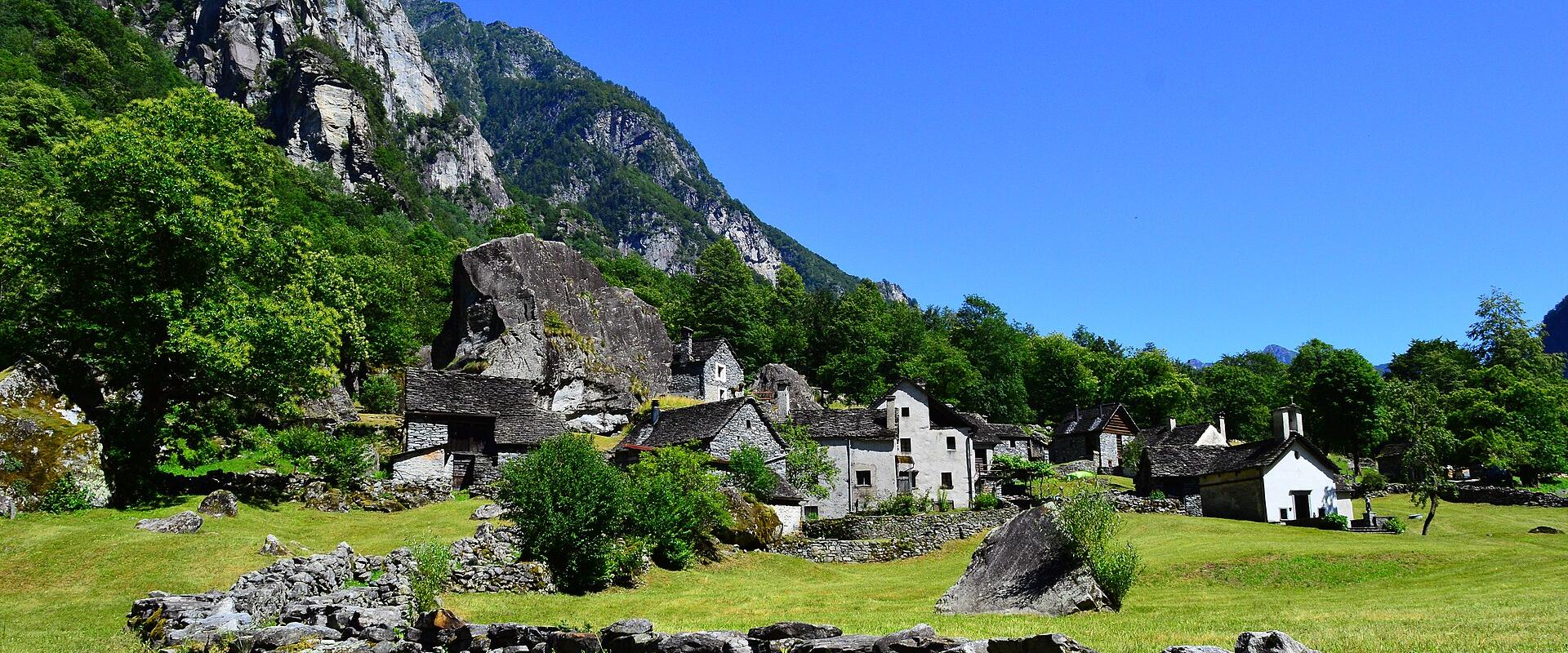 A landscape photograph showing a cluster of stone houses and a large boulder situated in a green valley at the foot of a steep, rocky mountain slope.