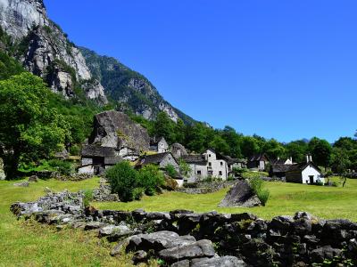 A landscape photograph showing a cluster of stone houses and a large boulder situated in a green valley at the foot of a steep, rocky mountain slope.