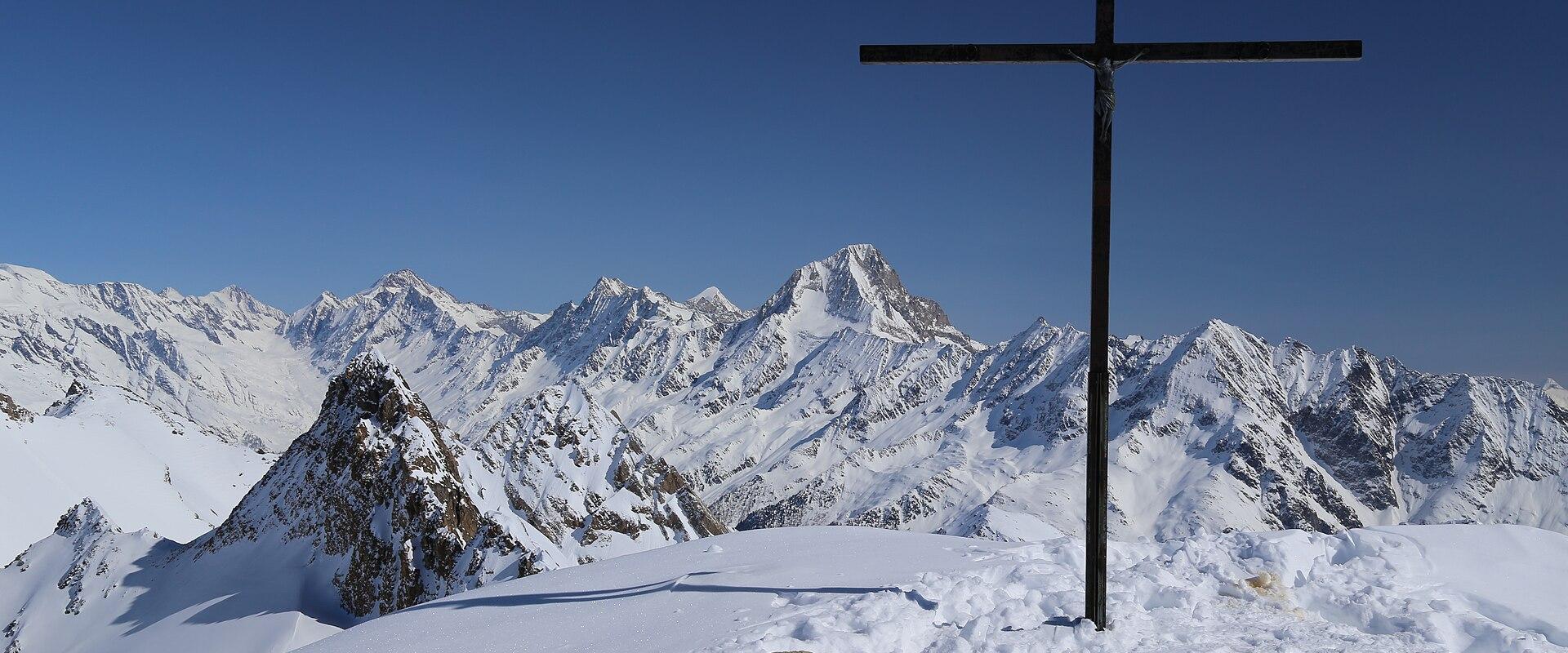 A large black cross stands on a snowy mountain summit in the foreground, overlooking a vast range of snow-covered alpine peaks under a clear blue sky.