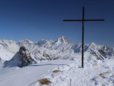 A large black cross stands on a snowy mountain summit in the foreground, overlooking a vast range of snow-covered alpine peaks under a clear blue sky.