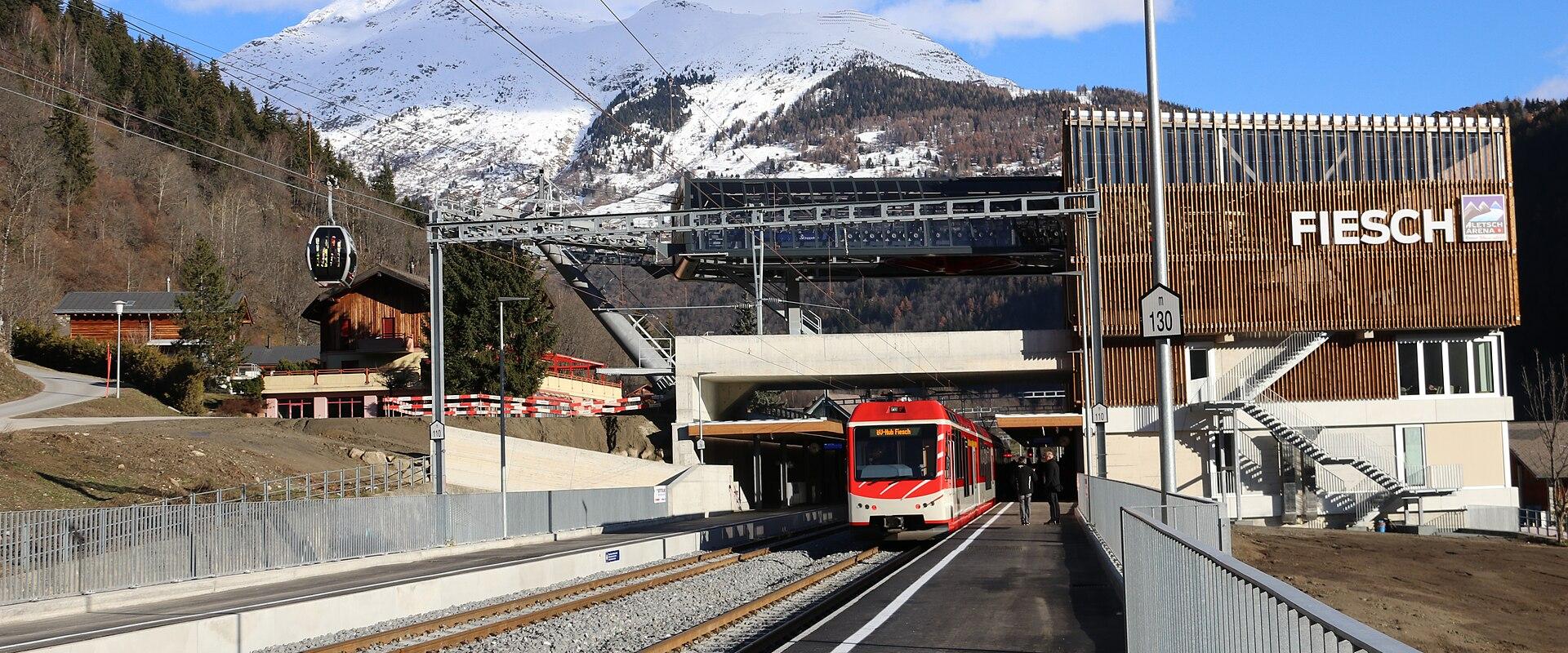 A red train at a railway station platform with a modern building on the right displaying the name "FIESCH" in large white letters, set against a backdrop of snow-capped mountains and a cable car.
