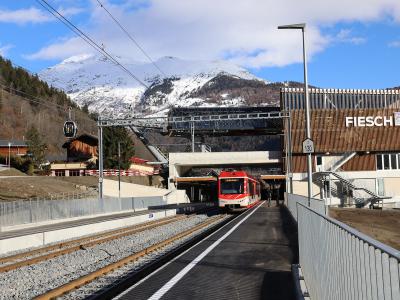 A red train at a railway station platform with a modern building on the right displaying the name "FIESCH" in large white letters, set against a backdrop of snow-capped mountains and a cable car.