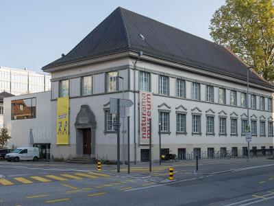 A street-level view of a large, light-colored building with a dark gabled roof, featuring a prominent vertical red banner reading "naturama museum" and a yellow exhibition banner on the facade, situated at a street corner with a crosswalk and parked 