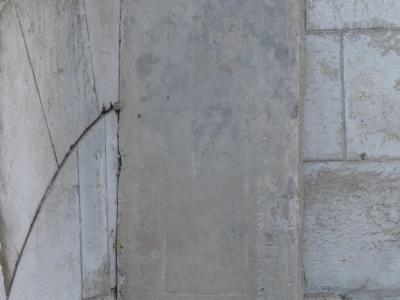 A weathered, upright stone slab or tombstone leaning against a corner of a light-colored stone wall, with a curved line etched into the adjacent wall section.