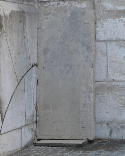 A weathered, upright stone slab or tombstone leaning against a corner of a light-colored stone wall, with a curved line etched into the adjacent wall section.
