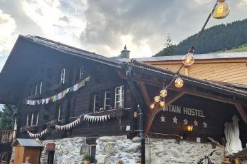 A traditional Swiss-style wooden chalet with a stone foundation, decorated with string lights and bunting flags, featuring a sign that reads "MOUNTAIN HOSTEL" with four stars beneath it, set against a backdrop of green hills and distant mountains