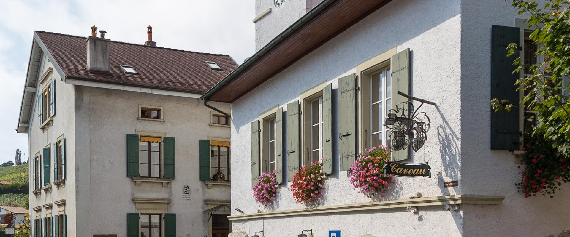 A street view of a white building with green shutters and flower boxes, featuring a sign for "Caveau" and a church tower with a clock in the background.