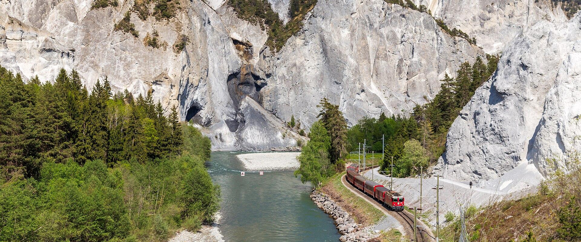 A red train travels along a railway track next to a river at the base of massive, light-colored limestone cliffs with visible rockfall debris.