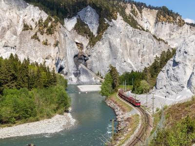 A red train travels along a railway track next to a river at the base of massive, light-colored limestone cliffs with visible rockfall debris.