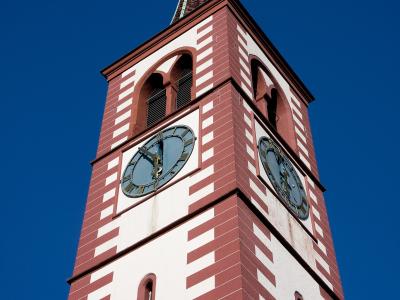 A low-angle shot of a tall church tower with red and white striped masonry, arched belfry openings, two clock faces, and a green spire topped with a weather vane against a clear blue sky.