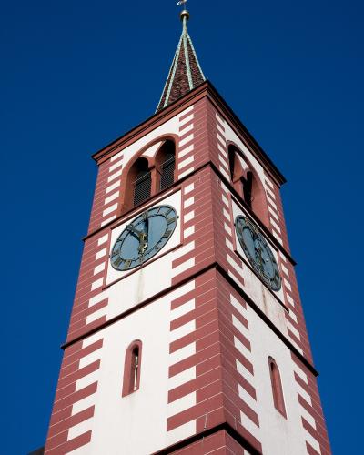 A low-angle shot of a tall church tower with red and white striped masonry, arched belfry openings, two clock faces, and a green spire topped with a weather vane against a clear blue sky.