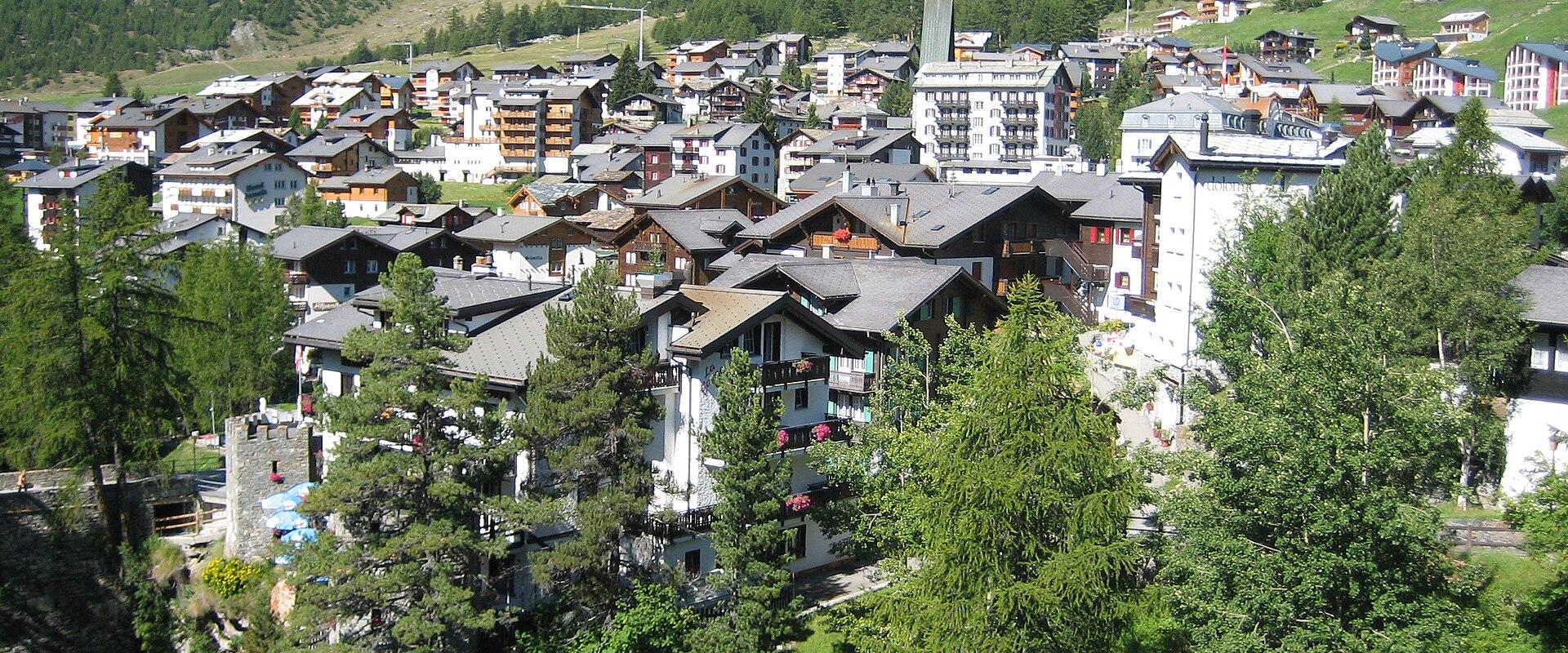 A high-angle view of a dense alpine village with traditional chalet-style architecture, surrounded by green slopes and a large mountain face in the background.