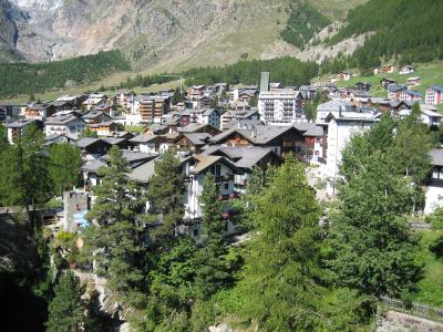 A high-angle view of a dense alpine village with traditional chalet-style architecture, surrounded by green slopes and a large mountain face in the background.