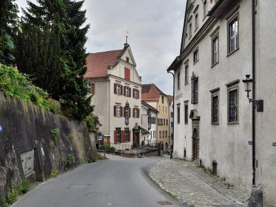 A narrow street scene in a historic European town showing a curved road, a retaining wall with vegetation on the left, and traditional buildings on the right, including one with a sign reading "MUSEUM KUNST" and a street sign for "Süsswinkelgasse".