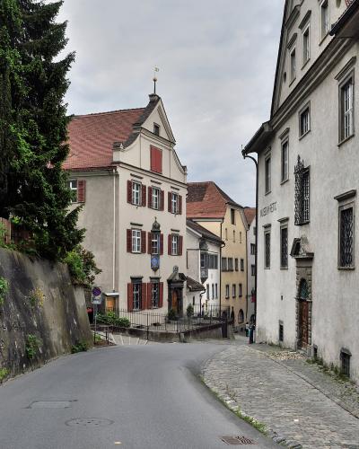 A narrow street scene in a historic European town showing a curved road, a retaining wall with vegetation on the left, and traditional buildings on the right, including one with a sign reading "MUSEUM KUNST" and a street sign for "Süsswinkelgasse".