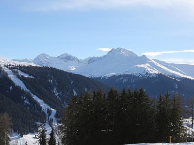 A snowy mountain landscape featuring a ski slope with visible tracks on the left, dense coniferous forest in the mid-ground, and a prominent snow-covered mountain peak in the background under a blue sky.