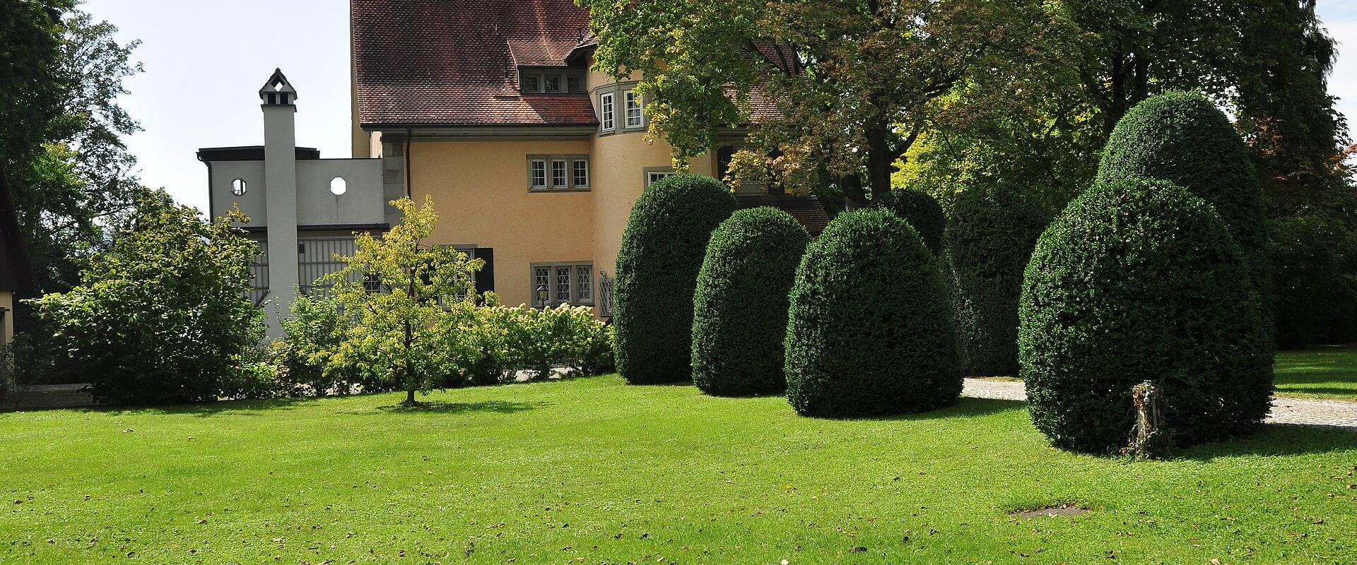 A yellow residential house with a red-tiled roof and a distinctive white chimney structure, viewed from a green lawn with manicured topiary bushes.