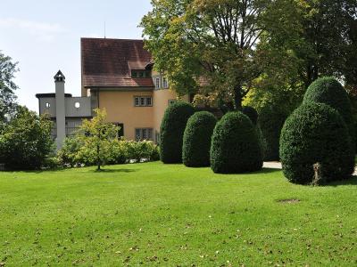 A yellow residential house with a red-tiled roof and a distinctive white chimney structure, viewed from a green lawn with manicured topiary bushes.
