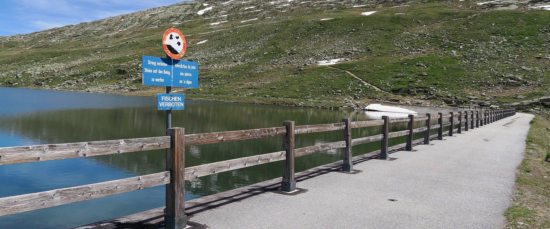 A paved path runs alongside a wooden fence bordering a calm alpine lake, with a blue signpost displaying German and French prohibitions against throwing trash and fishing, set against a grassy, snow-patched hillside.
