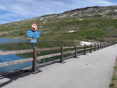 A paved path runs alongside a wooden fence bordering a calm alpine lake, with a blue signpost displaying German and French prohibitions against throwing trash and fishing, set against a grassy, snow-patched hillside.
