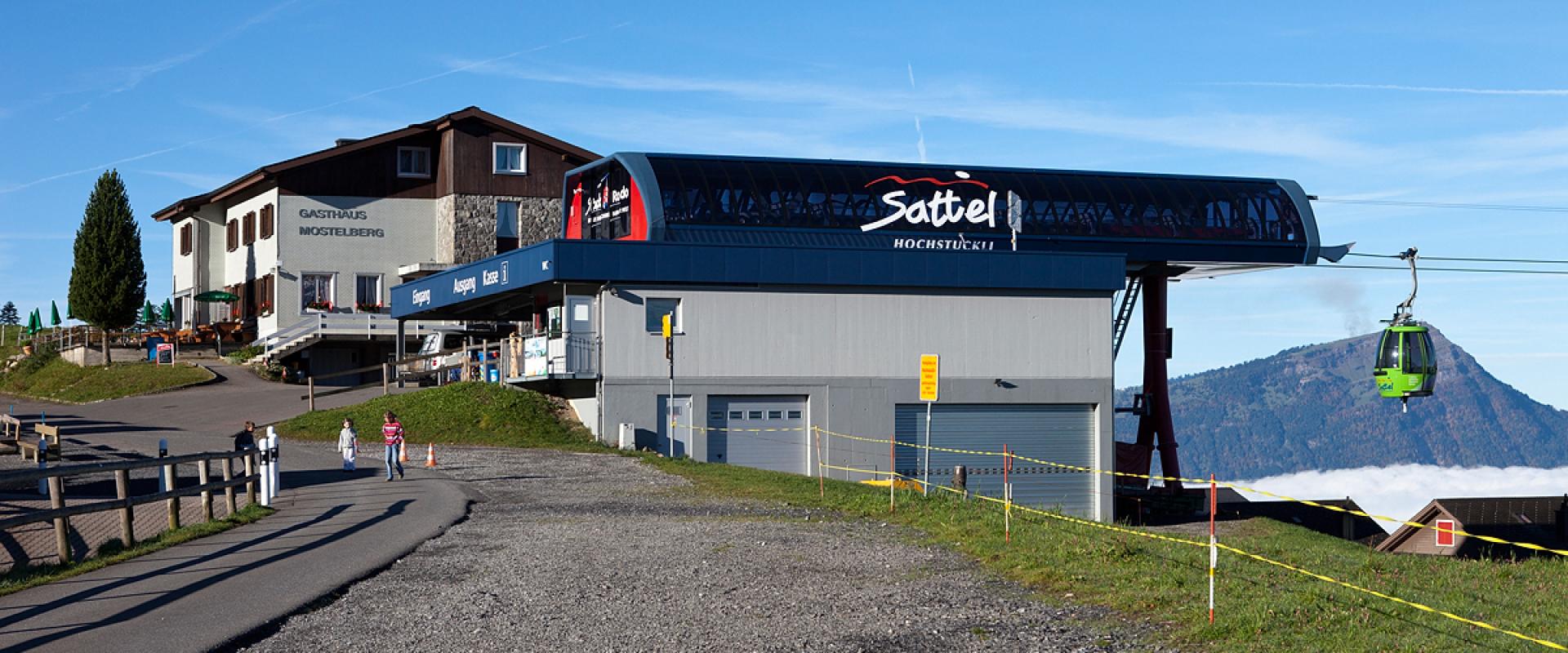 The image shows the upper station of a cable car system with a green gondola, a building labeled "GASTHAUS MOSTELBERG", and a large sign on the station roof reading "Sattel HOCHSTÜCKLI".