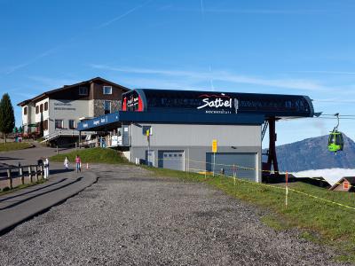 The image shows the upper station of a cable car system with a green gondola, a building labeled "GASTHAUS MOSTELBERG", and a large sign on the station roof reading "Sattel HOCHSTÜCKLI".