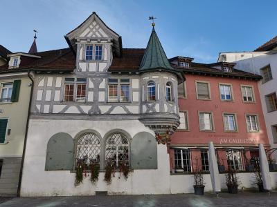 A street-level view of historic buildings in a European town square, featuring a white half-timbered house with a turret and a pink building with a sign reading "RESTAURANT & CAFE AM GALLUSPLATZ".