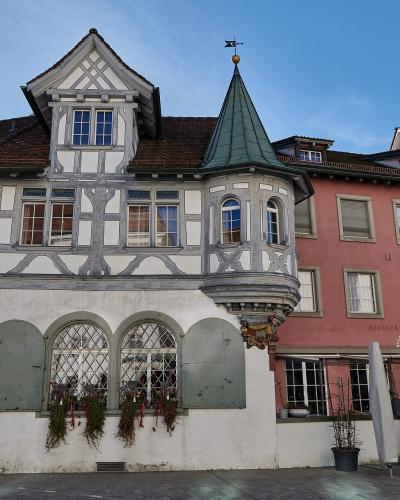 A street-level view of historic buildings in a European town square, featuring a white half-timbered house with a turret and a pink building with a sign reading "RESTAURANT & CAFE AM GALLUSPLATZ".