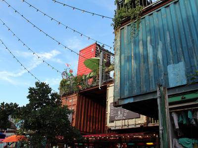 The image shows a multi-level outdoor structure constructed from stacked, repurposed shipping containers (blue, red, and brown), decorated with string lights, plants, and a bicycle on a balcony, under a blue sky.